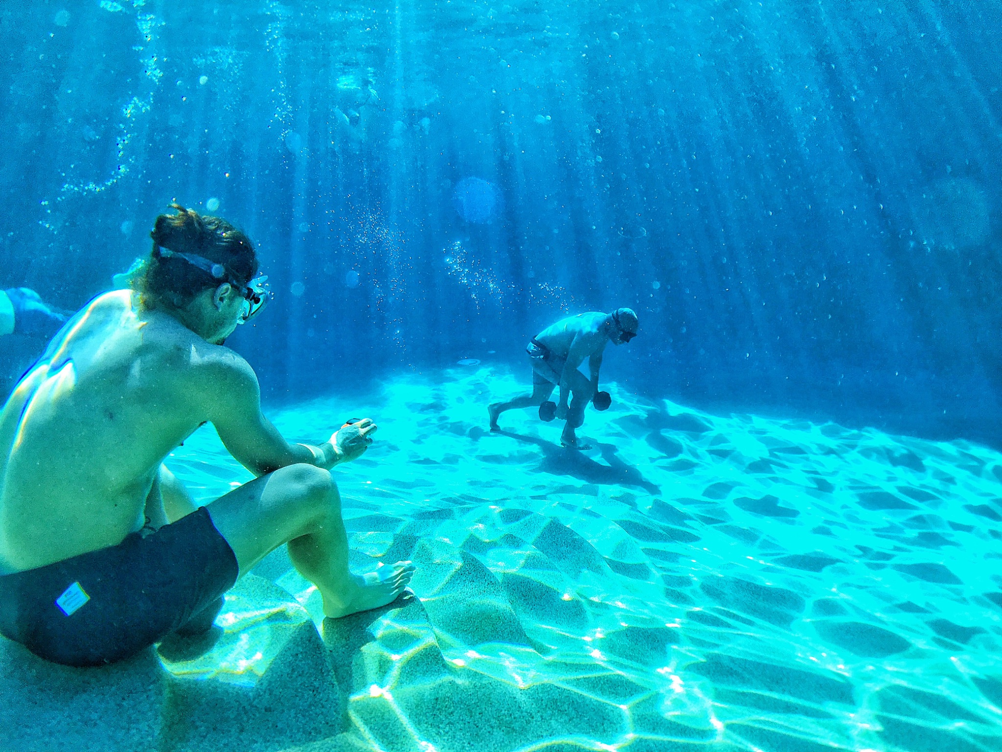 Two athletes doing underwater dumbbell training on the bottom of a clear blue pool